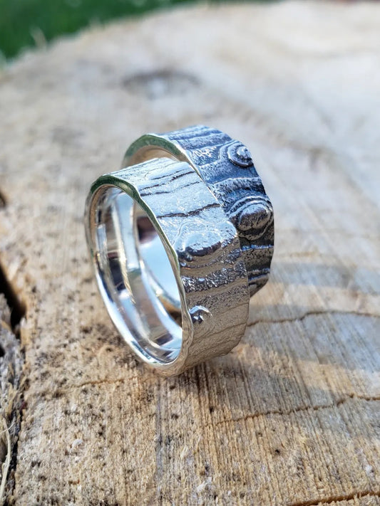 Two silver rings with bark texture, one polished and one oxidized.  They are sitting on their edges on a wooden surface.