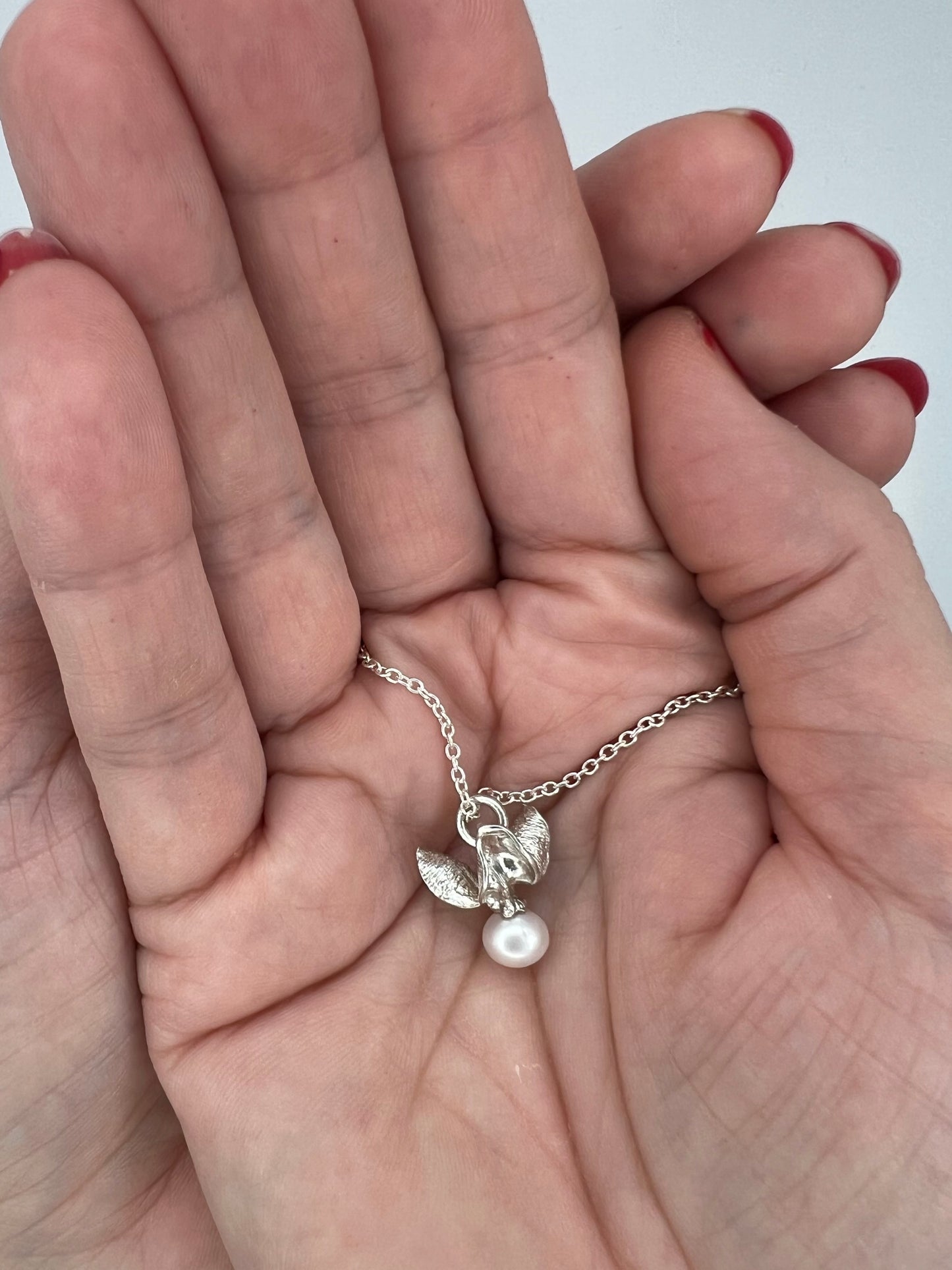 Two clasped hands holding a silver pendant with a white pearl on a fine cable chain. . The pendant is shaped like the shell & berry from a bittersweet vine. Hands are in front of a white background.