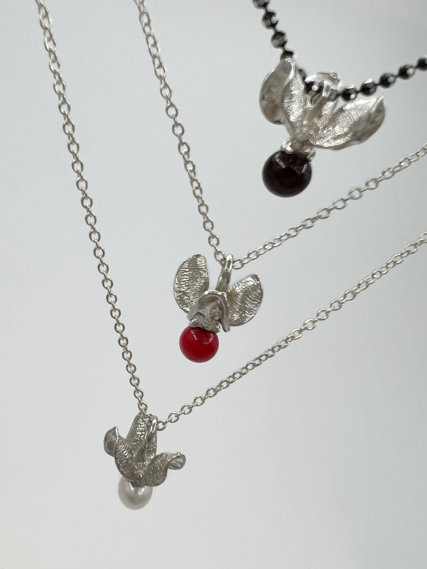 Three pendants hanging one above the other, depicted from an angle. They are silver with white pearl (bottom), red bamboo (middle) and black garnet (top) gem beads. The pendants are shaped like the shell & berry from a bittersweet vine. Shown against a white background.
