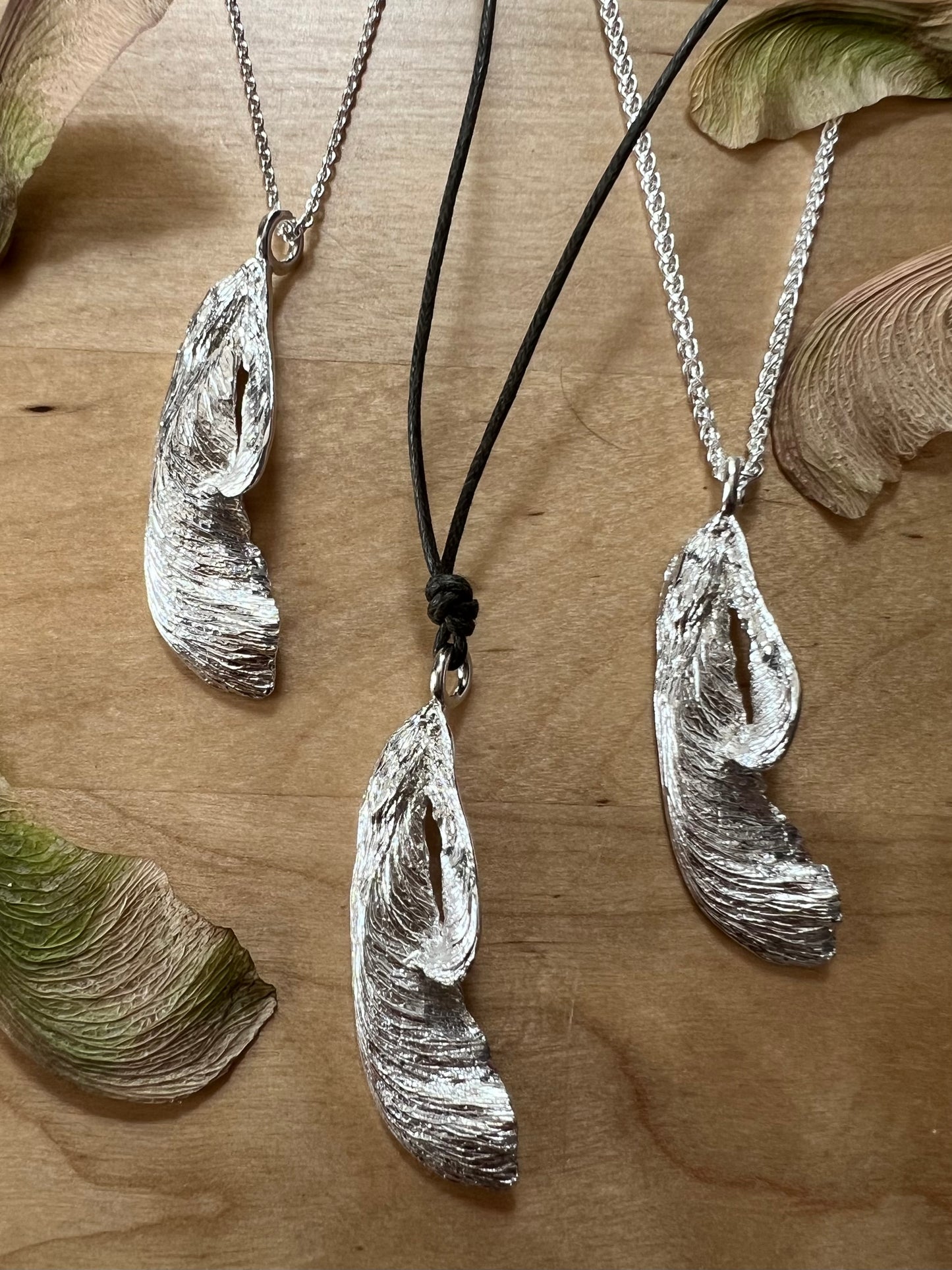Three silver maple key pendants displayed on a wooden table amongst real maple keys. The pendants are arranged in a "v" shape with the left-most on a fine chain, the centre on a green cord, and the right-most on a heavy chain.