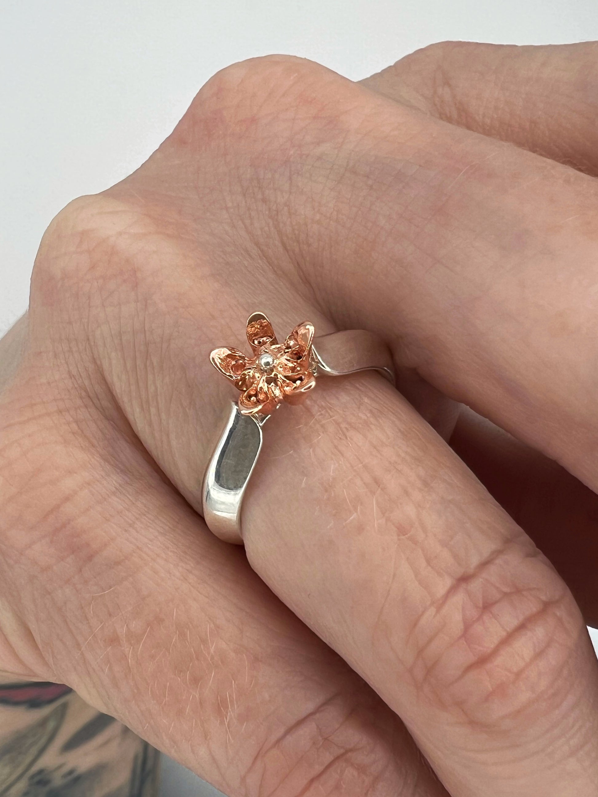 Hand displaying a ring with sterling silver band shaped like a leaf with a pink gold milkweed flower rising from between the "leaves"