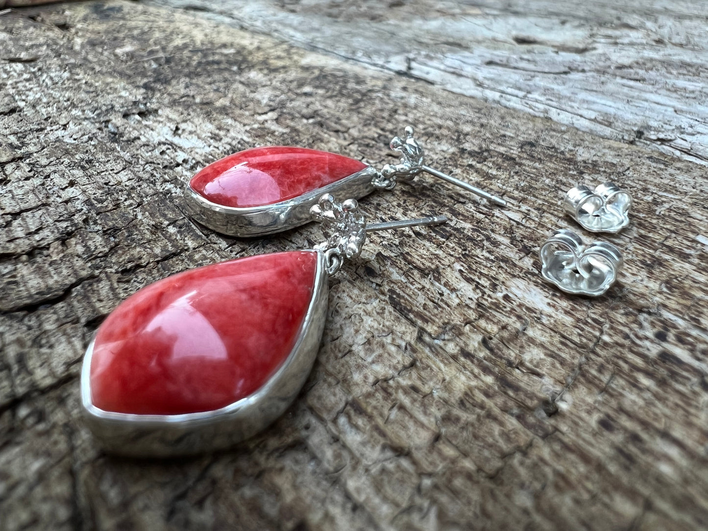 Silver earrings displayed on cracked wood. There are tiny crown shaped studs, beneath which dangle a larger coral-cocloured cabochon gem shaped like a teardrop. Shown from the side with the butterfly backs taken off the posts.
