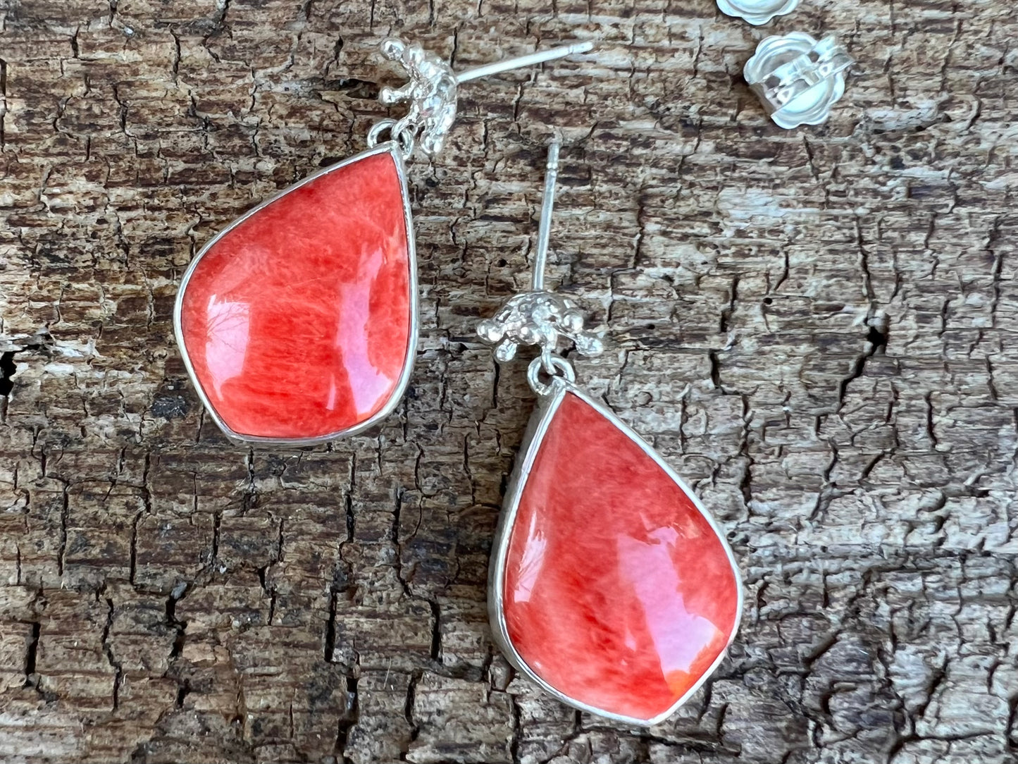 Silver earrings displayed on cracked wood. There are tiny crown shaped studs, beneath which dangle a larger coral-cocloured cabochon gem shaped like a teardrop. Shown with the butterfly backs taken off the posts.