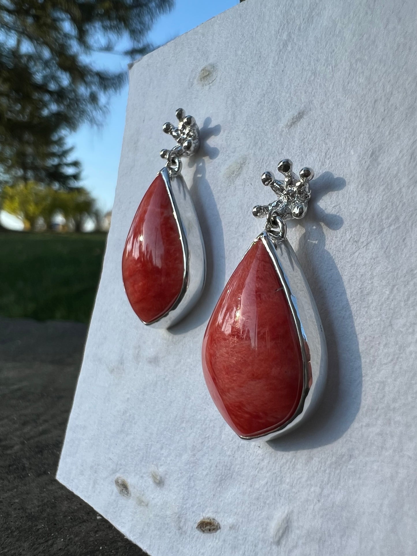 Silver earrings displayed on white seed paper. There are tiny crown shaped studs, beneath which dangle a larger coral-cocloured cabochon gem shaped like a teardrop. The card is positioned at an angle to show the sides of the earrings. Greenery & a tree can be seen in the background.