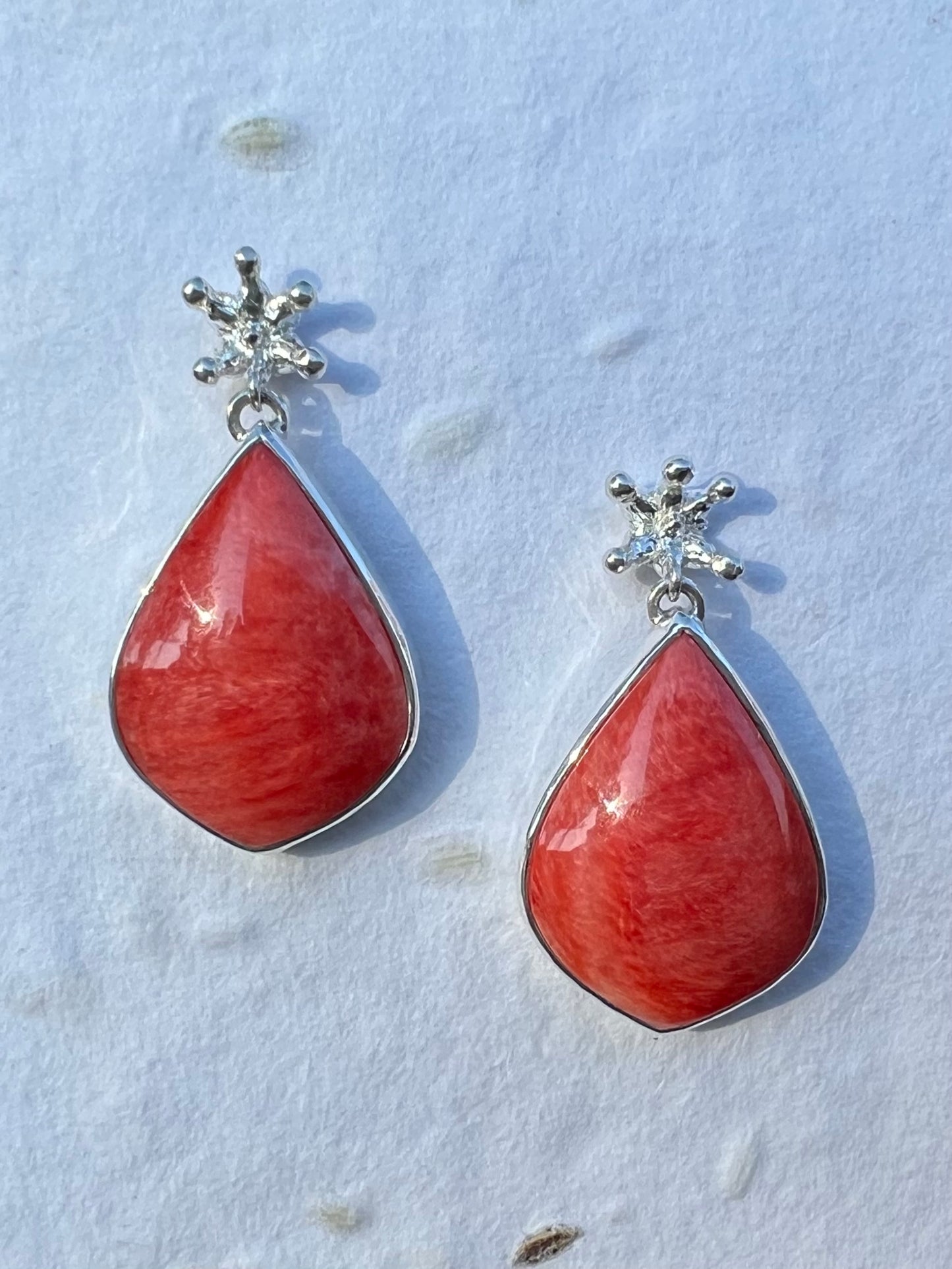Silver earrings displayed on white seed paper. There are tiny crown shaped studs, beneath which dangle a larger coral-cocloured cabochon gem shaped like a teardrop.