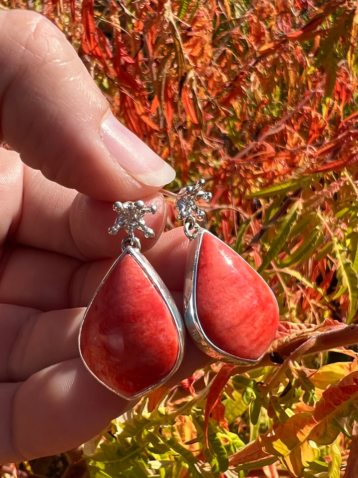 Fingers holding a pair of silver dangle earrings in front of orange fall foliage. The earrings are tiny crown shaped studs, beneath which dangle a larger coral-cocloured cabochon gem shaped like a teardrop.