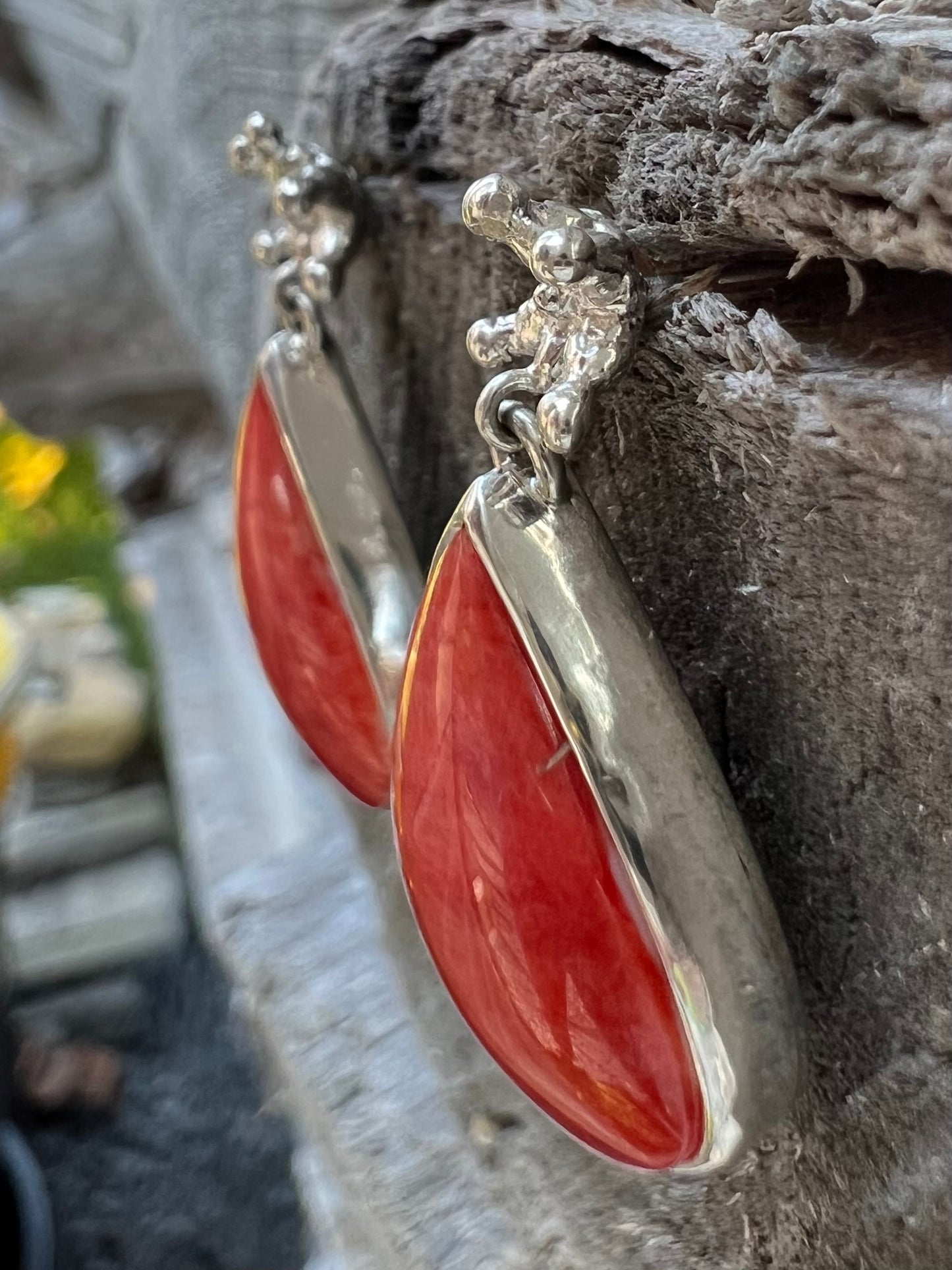 Silver earrings hanging from a wooden log. There are tiny crown shaped studs, beneath which dangle a larger coral-cocloured cabochon gem shaped like a teardrop. Photographed from an angle to show the side of the earring.