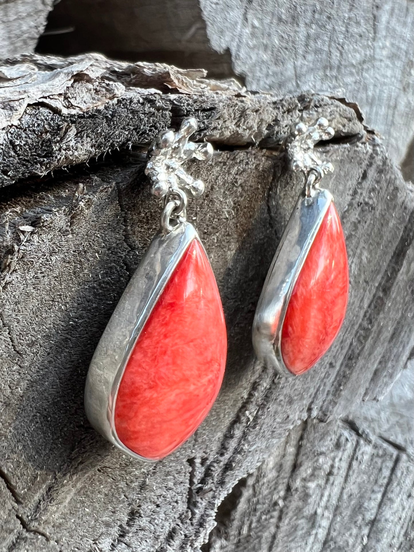 Silver earrings hanging from a wooden log. There are tiny crown shaped studs, beneath which dangle a larger coral-cocloured cabochon gem shaped like a teardrop.