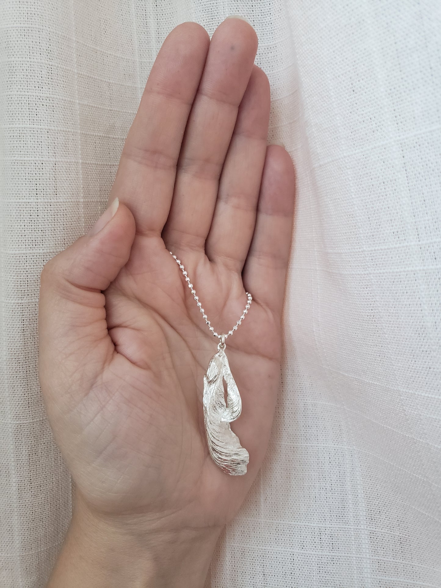 A silver maple key pendant a faceted ball chain in the palm of a hand.