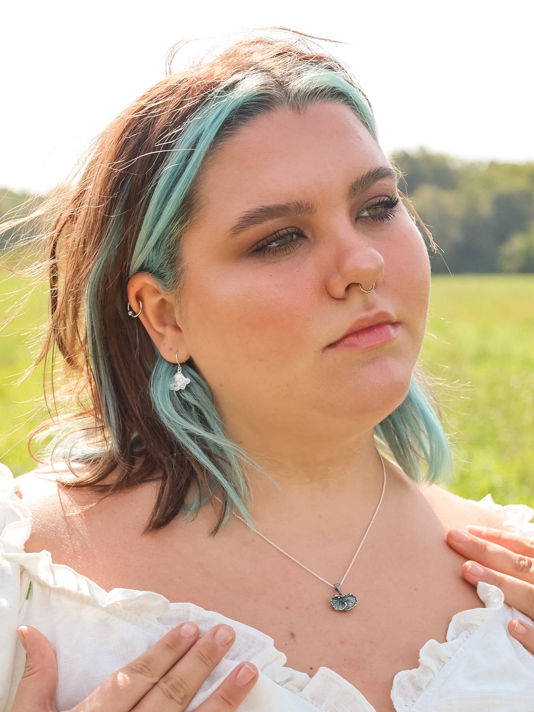 Woman in a field looking into the distance. She is wearing sterling silver fungi earrings, oxidized sterling silver fungi pendant and a white shirt.