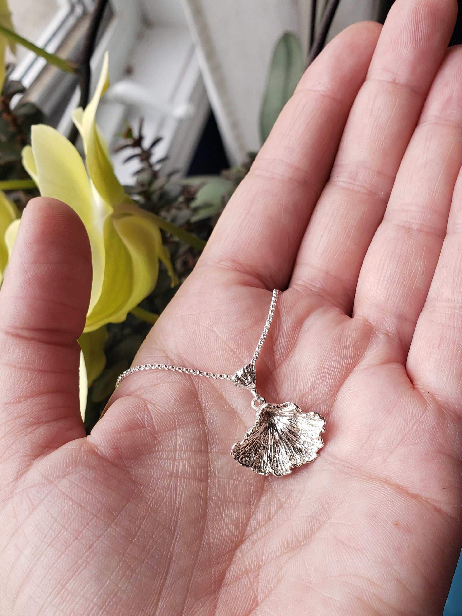 Hand holding a sterling silver fungi pendant with a ruffled edge. Yellow flowers are in the background.