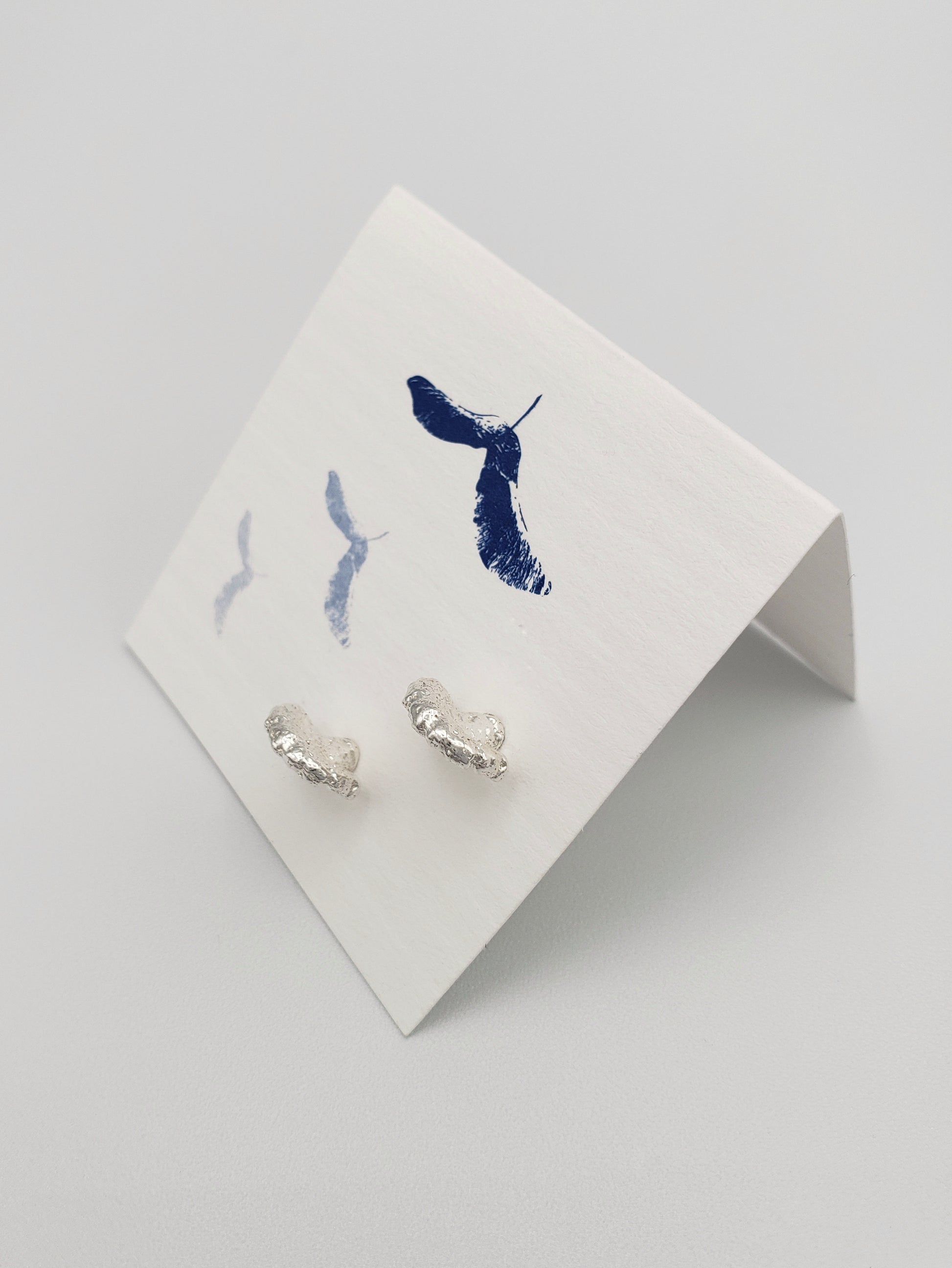 Sterling silver fungi stud earrings. Displayed on a white card with blue maple keys printed on it. Earrings stand out from the card as if they were growing from it.