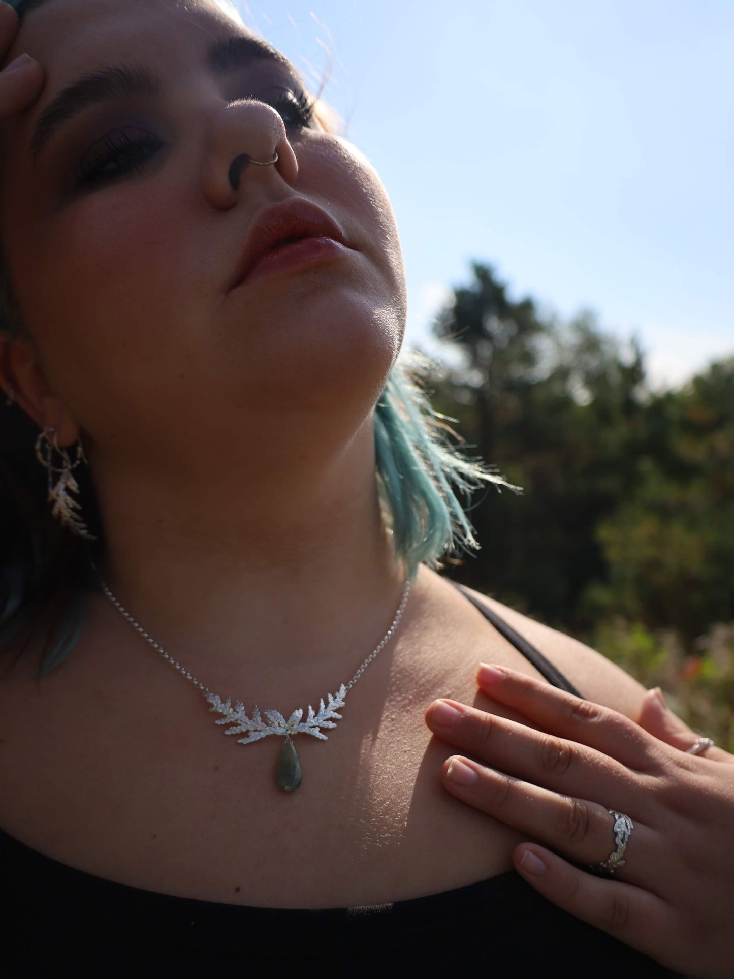 Woman standing in front of trees wearing sterling silver cedar jewellery / jewelry; including a large necklace with green sapphire briolette, cedar dangle and hoop earrings, and cedar rings.