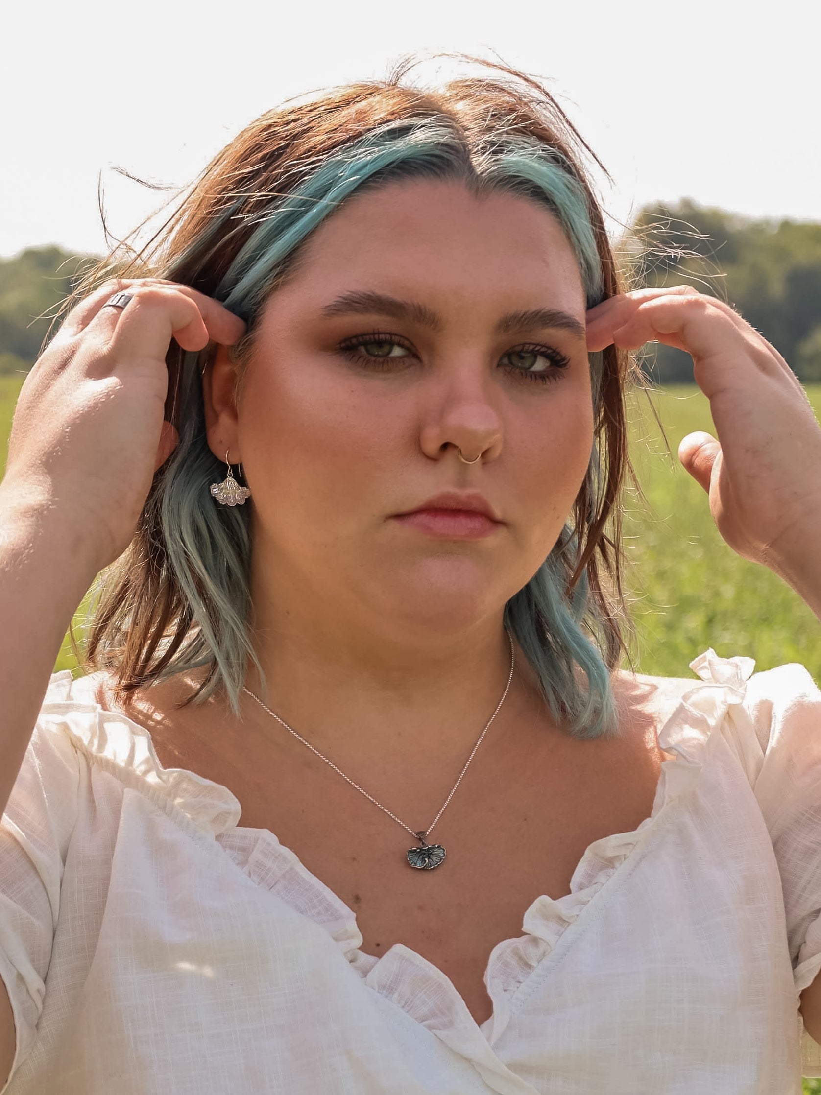 Woman in a field touching her hair. She is wearing sterling silver fungi earrings, oxidized sterling silver fungi pendant and a white shirt.