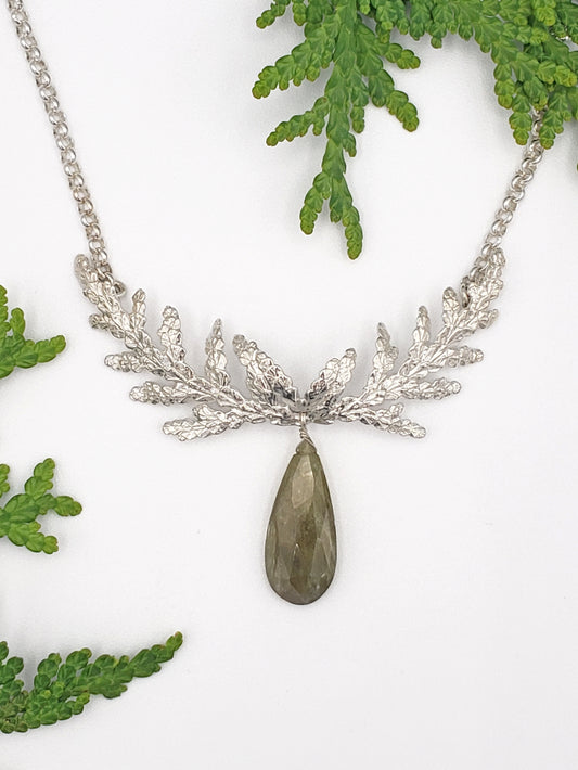 Sterling silver necklace made from cedar sprigs. Cedar is formed to resemble antlers and there is a large, tear-shaped green sapphire hanging from the bottom of the necklace. There are cedar sprigs in the background.