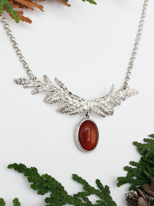 Sterling silver necklace made from cedar sprigs. Cedar is formed to resemble antlers and has a bezel set carnelian cabochon hanging from the bottom of the necklace. There are cedar sprigs in the background.