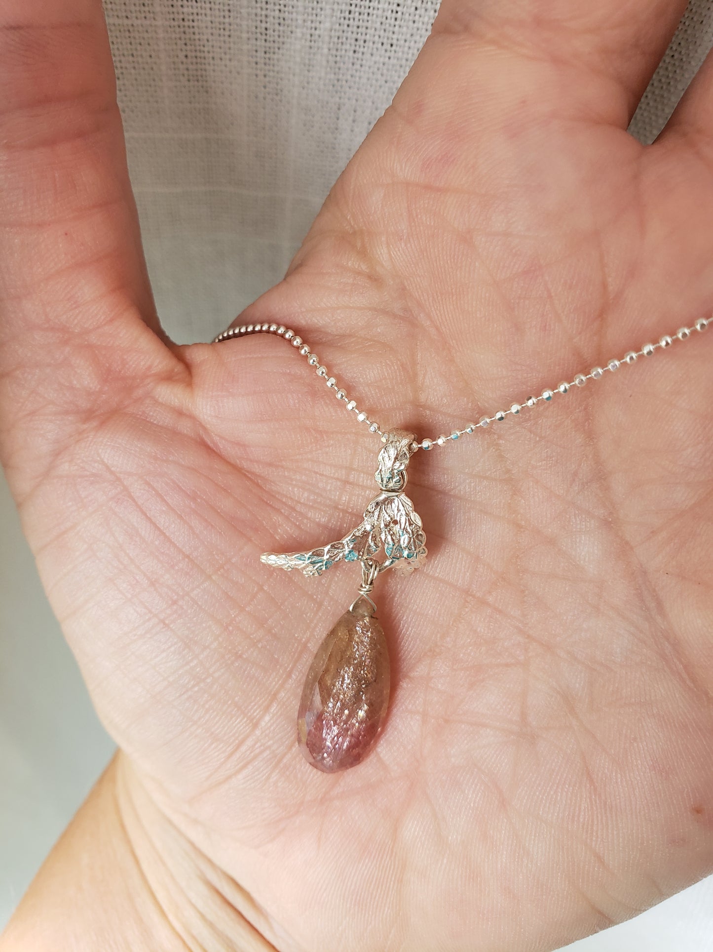 Hand holding a sterling silver cedar pendant with pink and tan umba sapphire dangling from the bottom.