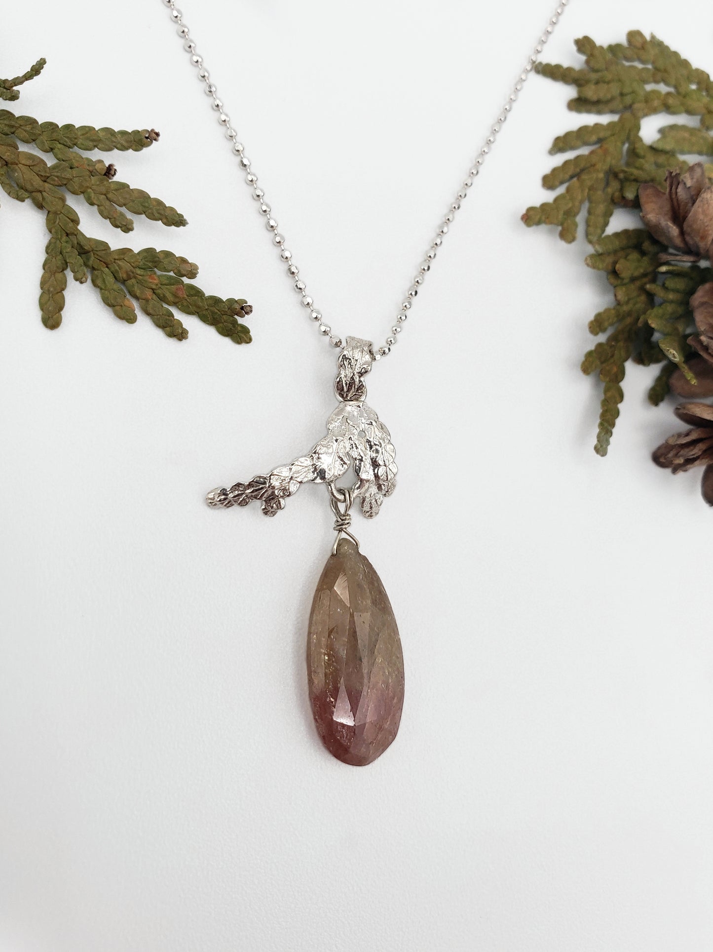 Sterling silver cedar pendant with pink and tan umba sapphire dangling from the bottom. There are cedar sprigs in the background.