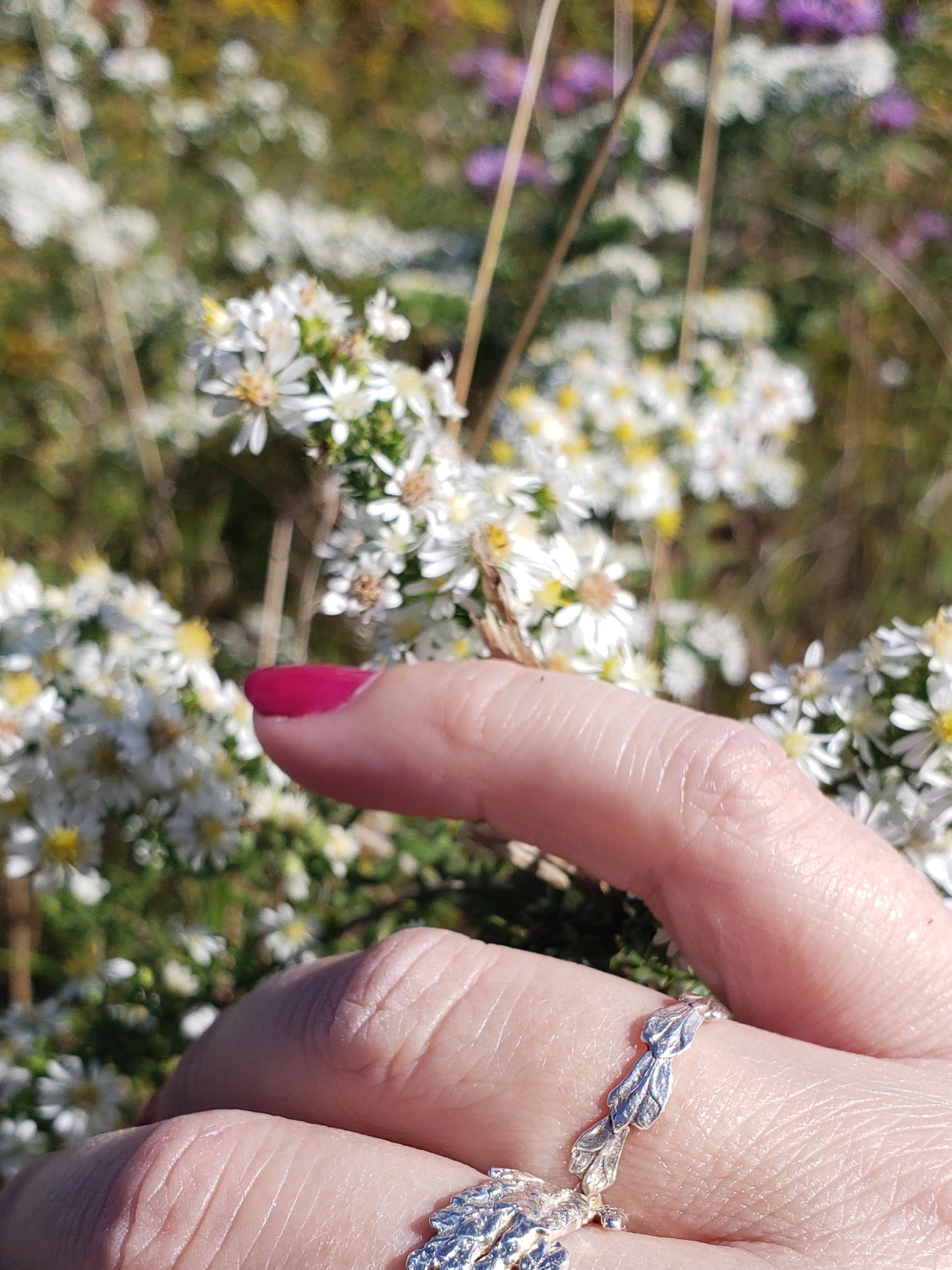 Hand wearing narrow sterling silver cedar ring with another ring out of view. There are white and purple flowers in the background.