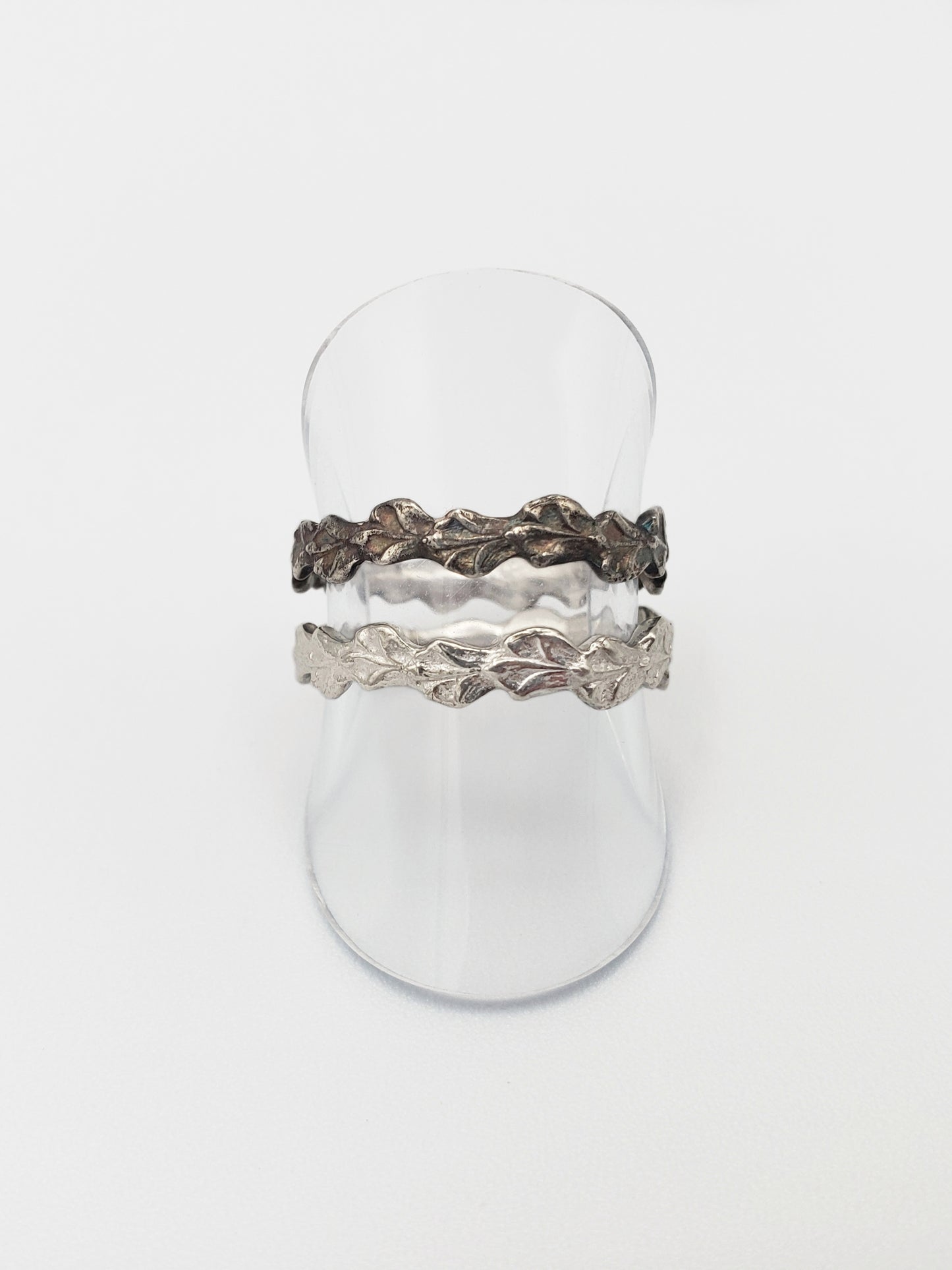 Two narrow sterling silver cedar rings displayed on a clear vinyl stand. The ring above is oxidized.